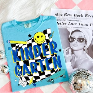 May include: A light blue t-shirt with the word "KINDERGARTEN" in blue and yellow lettering. The shirt features a checkered flag design, a smiley face, a pencil, a skateboard, and a skeleton hand making a peace sign.