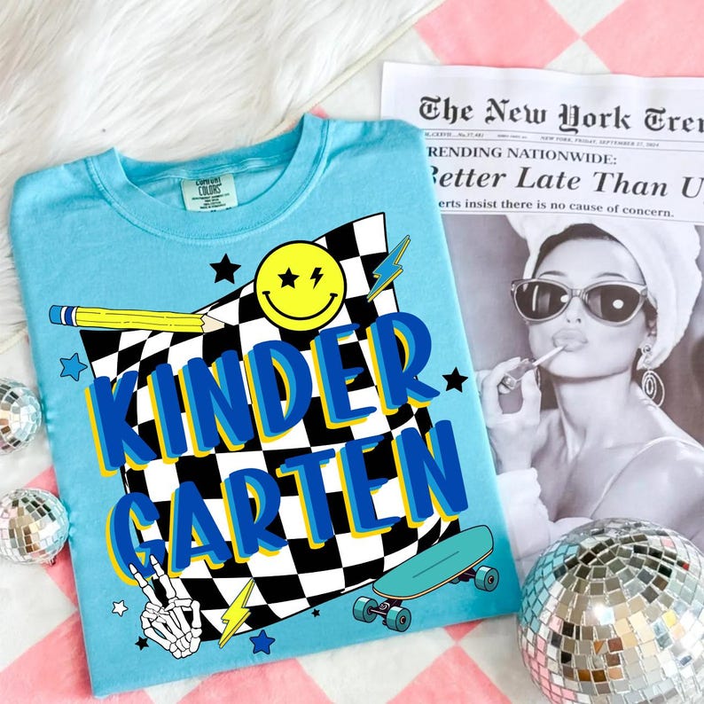 May include: A light blue t-shirt with the word "KINDERGARTEN" in blue and yellow lettering. The shirt features a checkered flag design, a smiley face, a pencil, a skateboard, and a skeleton hand making a peace sign.