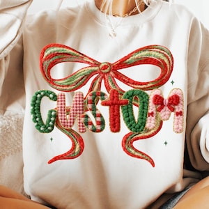 May include: Cream-colored sweatshirt with a large, decorative bow in red, green, and gold. The word "CUSTOM" is spelled out in a festive, textured font, with green, pink, and red elements. The design has a holiday theme.