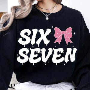 May include: Black sweatshirt with the words "SIX SEVEN" in white, dripping font. A pink bow graphic is on the right side. Small white star graphics are scattered around the text and bow.