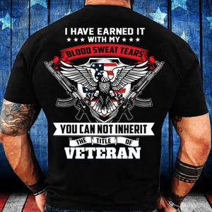 Può includere: T-shirt nero con grafica che include un'aquila, fucili e il testo "I HAVE EARNED IT WITH MY BLOOD SWEAT TEARS" e "YOU CAN NOT INHERIT THE TITLE OF VETERAN". Il design incorpora elementi rossi, bianchi e blu.