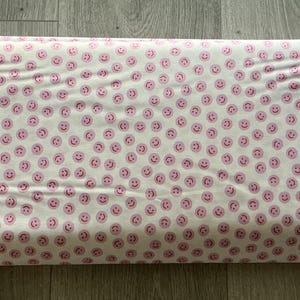 May include: A rectangular fabric roll featuring a white background with a repeating pattern of pink smiley faces. The edges of the fabric are a dark color. The fabric is folded and appears ready for use in sewing or crafting projects.