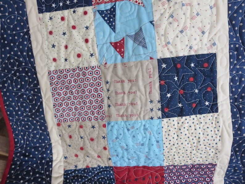 May include: A patchwork quilt with a patriotic theme. The quilt features squares of blue, red, and white fabrics with star, flag, and geometric patterns. Some squares have the text "Thank you!". The quilt is bordered with a blue fabric with white stars.