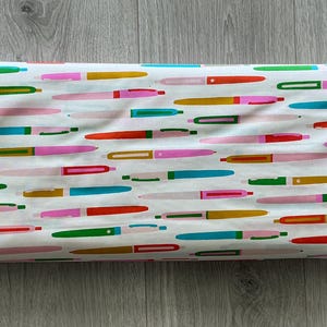 May include: A roll of white fabric with a colorful pen pattern. The pens are in various colors, including pink, green, yellow, orange, and blue. The pens are arranged horizontally across the fabric.