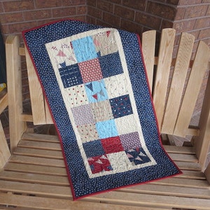 May include: A rectangular quilt with a patchwork design, featuring squares in various patterns and colors, including red, blue, and beige. The quilt has a navy blue border with white dots and a red binding. It is draped over a wooden chair.