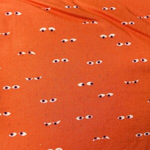May include: Orange fabric with a repeating pattern of white and purple-eyed, cartoon-style eyes. The fabric is folded, and a dark blue fabric with white text is visible on the left side.