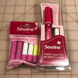 May include: Two packages of Sewline Fabric Glue Pens. One package contains a single pink pen with a butterfly design. The other package contains six glue pen refills in assorted colors: pink, white, and yellow. The text on the packages reads "Sewline Fabric Glue Pen" and "Sewline Fabric Glue Pen Refills".