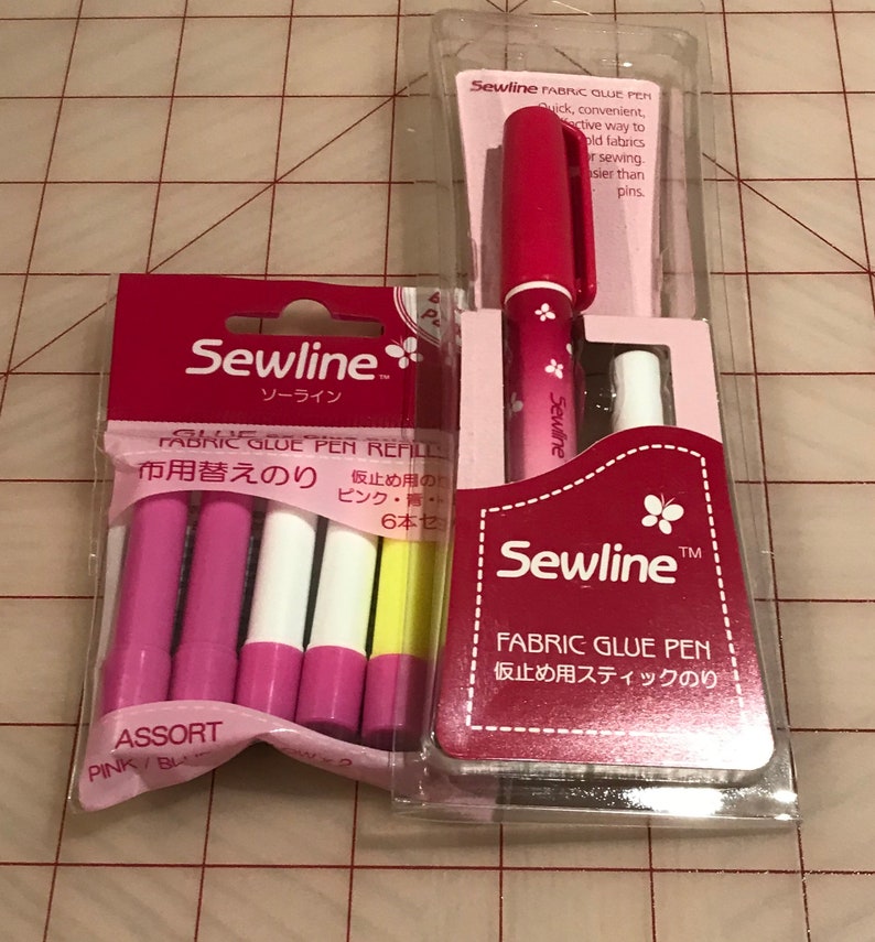 May include: Two packages of Sewline Fabric Glue Pens. One package contains a single pink pen with a butterfly design. The other package contains six glue pen refills in assorted colors: pink, white, and yellow. The text on the packages reads "Sewline Fabric Glue Pen" and "Sewline Fabric Glue Pen Refills".