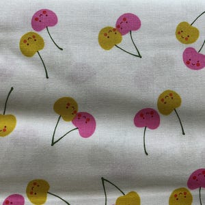 May include: White fabric with a repeating pattern of cartoon cherries. The cherries are yellow and pink with smiling faces and small green stems. The fabric has a slightly textured appearance.