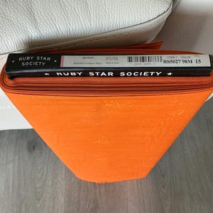May include: A roll of orange fabric with a speckled texture. The fabric is labeled "Ruby Star Society" with additional text and a barcode. The fabric is rolled up and appears to be new. The fabric is sitting on a white surface.