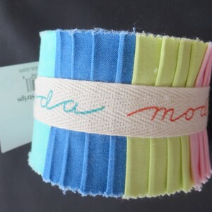 May include: A roll of fabric scraps in shades of blue, green, and pink. The fabric is folded and tied with a white ribbon that says "moda" in teal and orange.