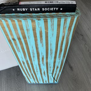 May include: A roll of fabric with a teal and gold vertical stripe pattern. The fabric is labeled "RUBY STAR SOCIETY" in black text. The roll is sitting on a wooden surface.
