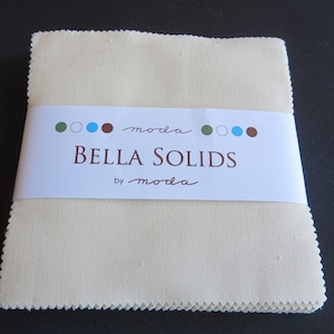 May include: A stack of cream-colored fabric squares, bound by a white label. The label reads "Bella Solids" in brown text, with the "moda" brand name above and below. The fabric has a slightly textured appearance.