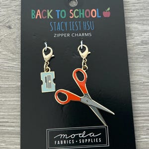 May include: Black card with two zipper charms. One charm is a light blue pencil sharpener, the other is a pair of red and gray scissors. The card reads "BACK TO SCHOOL STACY IEST HSU ZIPPER CHARMS" and "moda FABRICS + SUPPLIES".