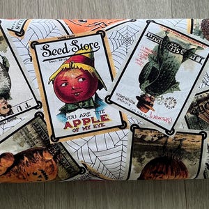 May include: Rolled fabric featuring vintage-style illustrations of scarecrows, pumpkins, and seed packets on a white background with spiderwebs. The fabric includes text that reads "You are the apple of my eye" and "Seed Store".