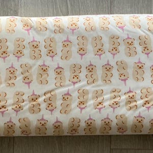 May include: A rolled fabric featuring a repeating pattern of light brown teddy bears with pink accents on a cream background. The bears are arranged in vertical rows. The fabric is likely cotton or a similar material.