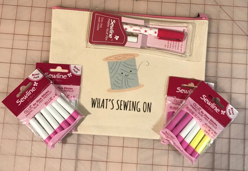 May include: A white canvas bag with a pink zipper and a cute cartoon spool of thread with the text "What's Sewing On". The bag contains two packs of Sewline glue sticks, one pack of blue glue sticks and one pack of assorted glue sticks. There is also a red and white fabric glue pen.