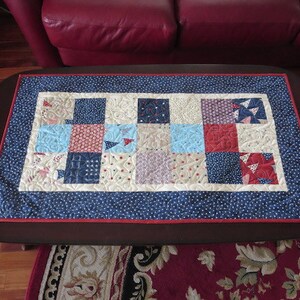 May include: A rectangular quilted table runner with a patchwork design. The center features squares in red, white, and blue patterns. A blue border with white dots surrounds the patchwork, with a thin red border. The runner is on a dark brown table.