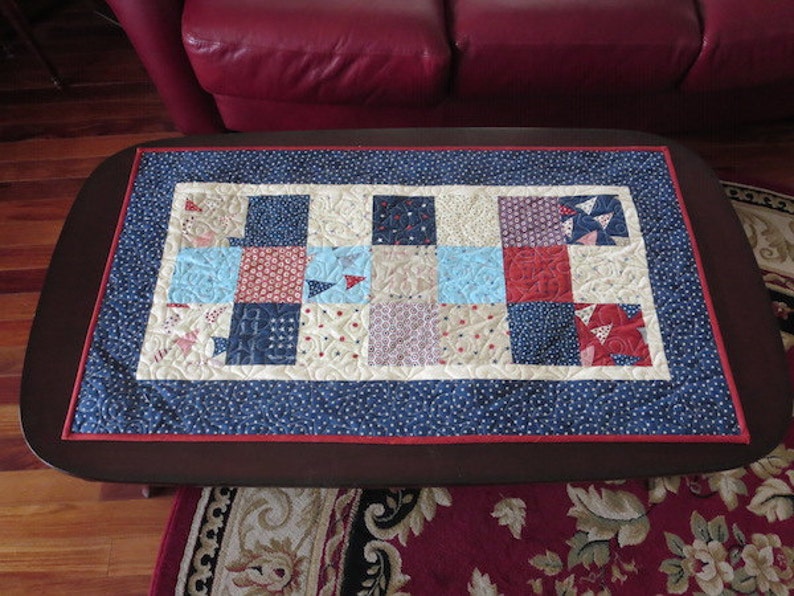 May include: A rectangular quilted table runner with a patchwork design. The center features squares in red, white, and blue patterns. A blue border with white dots surrounds the patchwork, with a thin red border. The runner is on a dark brown table.