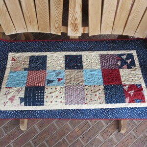May include: A rectangular quilted bench pad with a navy blue border featuring white stars and a red trim. The center has a patchwork design with squares in red, light blue, navy, and cream, with various patterns. The pad is placed on a wooden bench.