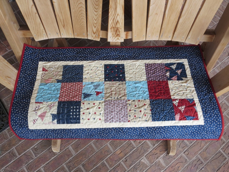 May include: A rectangular quilted bench pad with a navy blue border featuring white stars and a red trim. The center has a patchwork design with squares in red, light blue, navy, and cream, with various patterns. The pad is placed on a wooden bench.