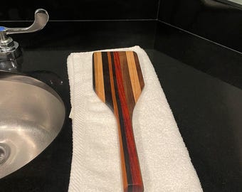 May include: A wooden spatula with a rectangular head and a long handle. The spatula is made of various wood tones, including brown, red, and black stripes. It rests on a white towel on a black countertop.