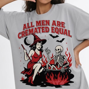 以下が含まれることがあります： 赤色のドレスを着た魔女と炎のある大釜の中にいる骸骨が描かれた、ライトグレーのTシャツです。「ALL MEN ARE CREMATED EQUAL」の文字がイラストの上にあります。2匹のコウモリと三日月も含まれています。