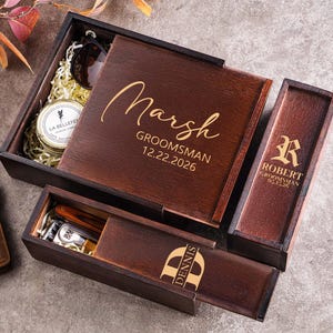 以下が含まれることがあります： 「Marsh」と「Groomsman 12.22.2026」という名前が蓋に刻まれた、濃い色の木製ギフトボックスセット。ボックスにはサングラス、キャンドル、その他のアイテムが入っています。名前と日付が記載された追加のボックスも含まれています。