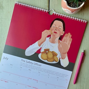以下が含まれることがあります： 7月の壁掛けカレンダー「The Safety Officer」は、皿の上のクッキーを食べながら片手を上げている人物のグラフィックイラストが特徴です。カレンダーには日付のグリッドと手書きのメモが含まれています。