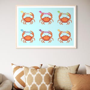 Puede incluir: Una impresión artística enmarcada con seis cangrejos de dibujos animados sobre un fondo azul claro. Cada cangrejo naranja tiene una diadema colorida con texto. La impresión se muestra sobre un sofá con cojines decorativos en tonos marrones y blancos.