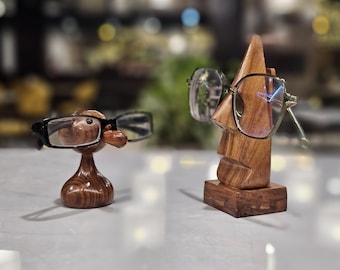 May include: Two wooden eyeglass holders. One is a small, rounded, smooth brown wooden figure with black-framed glasses. The other is a taller, triangular, carved wooden face with a pointed top and a rectangular base, wearing metal-framed glasses.