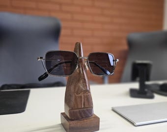 May include: A pair of square-framed sunglasses with dark lenses and a thin metallic frame, resting on a carved wooden nose-shaped display stand. The stand features a natural wood grain texture and a rectangular base, supporting the eyewear.