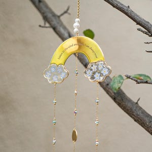 May include: A decorative wind chime featuring a yellow rainbow design with the text "Listen to the wind and I am always near." The rainbow has cloud accents with small white and blue flowers. The chime hangs from a gold chain with pearl and bead accents.