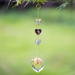 May include: A hanging heart-shaped wind chime with a gold chain and accents. The chime features three heart-shaped pendants with floral and pet photo inserts, and a clear crystal heart at the bottom. The bottom heart has the name "LILY" and a paw print.