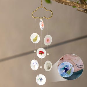 May include: A handmade wind chime featuring a gold-colored cloud-shaped top and a series of clear, circular pendants. Each pendant showcases pressed flowers and botanical elements. The wind chime is suspended from a tree branch.