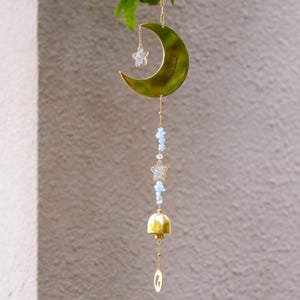 Puede incluir: Un carillón de viento dorado con un diseño de luna creciente y estrella. La luna tiene el texto "Always loved" grabado. El carillón incluye cuentas azules, una campana y un pequeño dije. Cuelga de una cadena dorada.