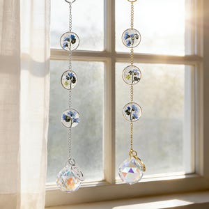 May include: Two decorative suncatchers hanging in front of a window. One has a silver chain and the other a gold chain. Each suncatcher features three circular pendants with blue flowers and a crystal ball at the bottom.