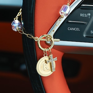 Op de afbeelding: Goudkleurige auto-hanger met een kruisvormige hanger versierd met heldere kristallen en een ronde hanger met de letter "K". De hanger hangt aan een ketting met iriserende kristallen kralen. De achtergrond toont het interieur van een auto.