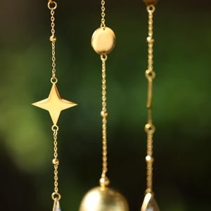 以下が含まれることがあります： さまざまな形の金色の風鈴のクローズアップ。風鈴は、星、丸いディスク、繊細なチェーンから吊り下げられた小さなビーズが特徴です。背景はぼやけた緑色で、光沢のある金との柔らかなコントラストを生み出しています。