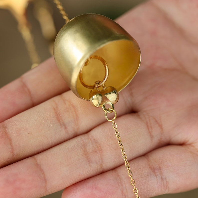 以下が含まれることがあります： 金色のベルペンダントのクローズアップ。ベルは丸みを帯びた形状で、小さな舌があり、繊細なゴールドチェーンから吊り下げられています。ベルは人の手で持たれています。