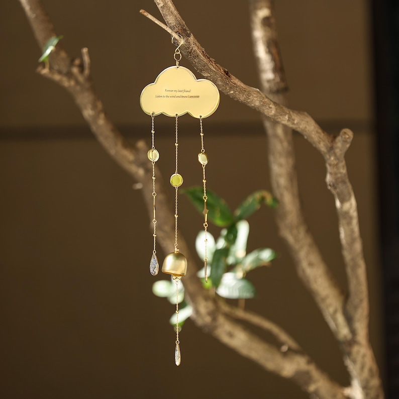以下が含まれることがあります： 雲の形をしたトップが付いた金色の風鈴で、木の枝から吊り下げられています。「Forever my best friend, Listen to the wind and know I am here」というテキストが書かれた雲。風鈴には、ビーズとベルが付いたぶら下がっているチェーンが付いています。