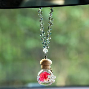 May include: A car air freshener with a clear glass bottle containing red petals, topped with a wooden cap. A beaded strand of iridescent beads hangs from the top. A white flower charm dangles from the bottle.
