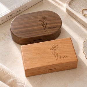 May include: Two wooden boxes with lids. The top oval box is dark brown wood with an engraved daffodil and the name Deanna. The bottom rectangular box is light brown wood with an engraved pansy flower and the name Sophia. Both feature a small slot.