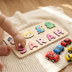 Könnte beinhalten: Ein Holzpuzzle mit dem Namen "SARAH" in rosa Buchstaben. Das Puzzle enthält tierförmige Teile: eine Katze, einen Papagei und eine Schildkröte. Kleine Spielzeugautos in Rot, Blau und Gelb sind in der Nähe angeordnet. Das Puzzle liegt auf einer cremefarbenen Decke.