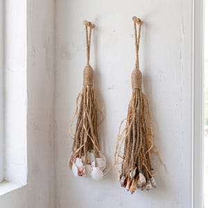 May include: Two hanging decorative tassels made of thick tan jute rope, each featuring a wrapped top loop and a bundle of loose, frayed fibers. The bottom of each tassel is adorned with a cluster of various white and brown seashells.