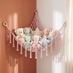 Op de afbeelding: Een roze macramé hoekhangmat voor speelgoed met een geknoopt netontwerp en hangende kwastjes langs de onderrand. De hangmat wordt opgehangen aan drie houten ringen en bevat een verzameling pluche knuffeldieren, waaronder beren en konijnen.