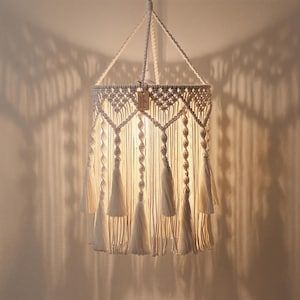 May include: A cream-colored macrame pendant light fixture featuring intricate knotted patterns, vertical fringe strands, and multiple tassels hanging from a circular frame. A small rectangular wooden tag is attached to the top structure.