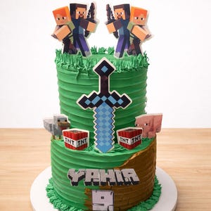 May include: A two-tiered green frosted cake decorated with Minecraft-themed elements. The cake features character figurines, a pixelated sword, TNT blocks, and the name "YAHIA" with the number "9". The cake sits on a white cake board.