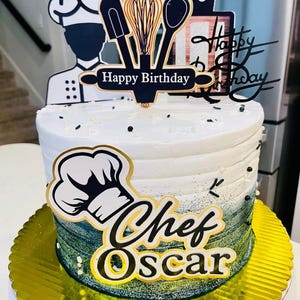 May include: A white frosted birthday cake with a chef theme. The cake has a gold base and is decorated with black and gold toppers that say "Happy Birthday" and "Chef Oscar". The cake is decorated with black sprinkles.
