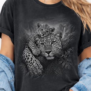 Comfort Colors® Pepper shirt featuring a detailed black and white leopard graphic with a leopard cub resting on top of an adult leopard in a jungle setting. Moody wildlife design with tropical leaves, rocky landscape, and realistic big cat artwork. Great animal graphic tee for leopard lovers, wildlife fans, safari style outfits, nature lovers, and unique animal gift ideas.